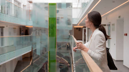 young business woman in a white jacket is standing in a public space with a takeaway coffee in her hand. Confident brunette freelancer, portrait on the background of a business center.の写真素材
