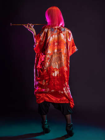 Futuristic look, multicultural outfit, young woman with pink hair in modern trousers, vintage kimono and smoking a pipe in her handsの写真素材