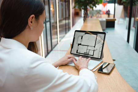 architect or interior designer works remotely. young freelance woman works with an engineering floor plan. brunette in a white jacket with a laptop in public space, drawing of apartment is on screen.の写真素材