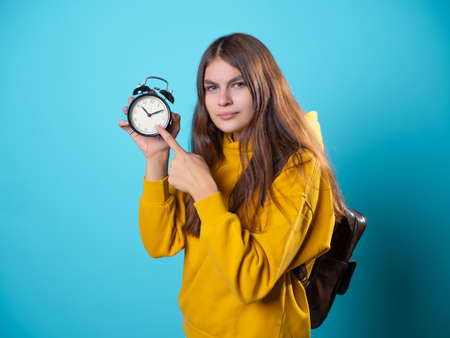 Hurry up, concept. A young brunette in a youth hoodie holds an alarm clock in her hands and points to the dial. A call to action, a bright photo on a blue background.の写真素材