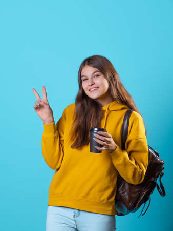 Cute young brunette in jeans and yellow hoodie. smiling student in glasses with a backpack on her shoulders holds a takeaway coffee in her hands, and shows a two thumbs up gestureの写真素材