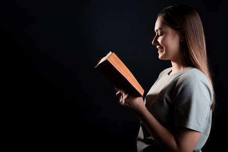 Reading a book, studying or enjoying a novel. Young brunette in a T-shirt with an open book in her hands, portrait in profile on a black backgroundの写真素材