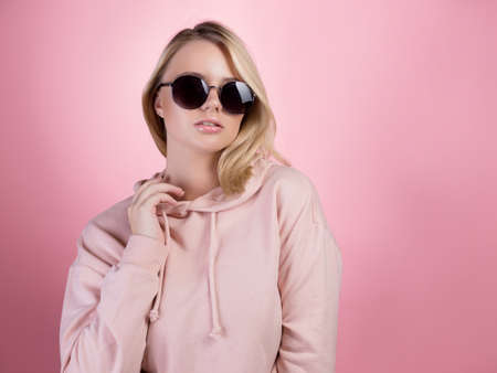 beautiful young woman with blonde hair wearing dark sunglasses. In a pink hoodie on a pink background, studio portraitの写真素材
