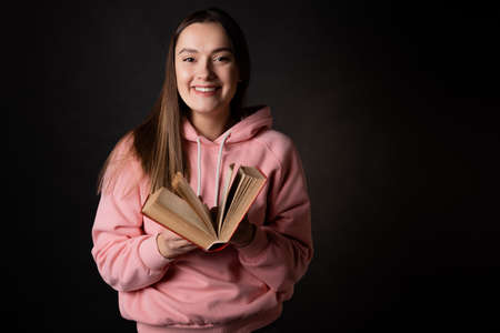 A young cute brunette in a pink hoodie, reading a book. Student with an open book in her hands, portrait on black. Study or read fictionの写真素材