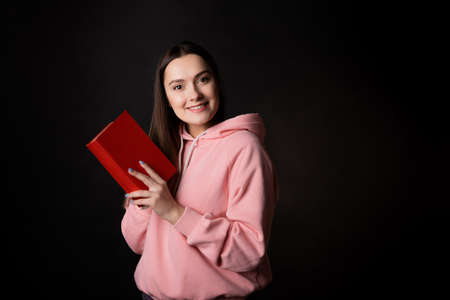 A young cute brunette in a pink hoodie, reading a book. Student with an open book in her hands, portrait on black. Study or read fictionの写真素材