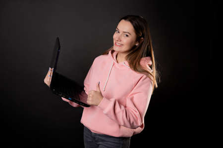 A happy student or freelancer with a laptop in her hands. A young charming woman in a pink hoodie works remotely and gives a thumbs up. Brunette holds an open laptop, photo on blackの写真素材