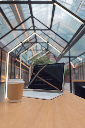 freelancers workplace. A thin mobile laptop on a table in a coworking or other public space. low depth of field. tablet computer with a code on the screen and coffee to take away. Copy space in frontの写真素材