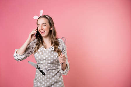 Young charming woman pastry chef communicates with customers by phone, take orders, entrepreneur. A cook in a polka-dot apron. pastry chef in an apron on a pink background.の写真素材