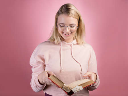 young female student, a portrait with an open book in her hands. Study and gain knowledge, preparation for admission to college or university, exams. Cute blonde with glasses and a pink hoodie.の写真素材