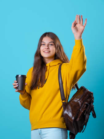 Cute young brunette in jeans and yellow hoodie. smiling student in glasses with a backpack on her shoulders holds a takeaway coffee in her hands and waves a hand in greeting, blue background.の写真素材