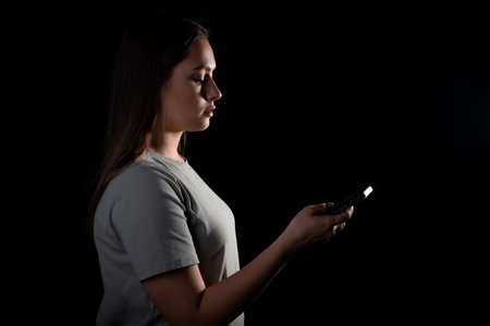 A calm portrait of a young brunette with a phone in her hands in profile. Contour light, copy the space on the right, photo on a black backgroundの写真素材