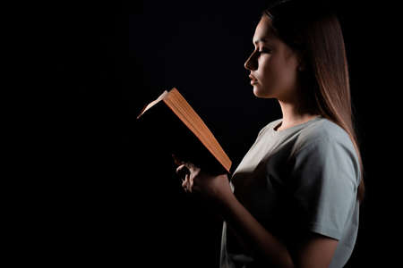 Reading a book, studying or enjoying a novel. Young brunette in a T-shirt with an open book in her hands, portrait in profile on a black backgroundの写真素材