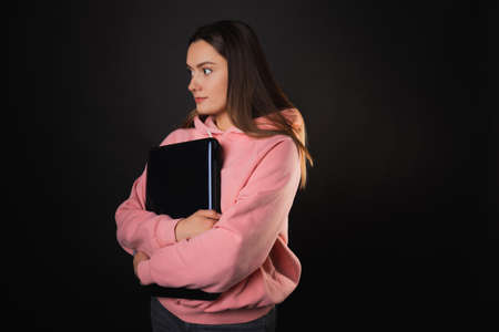 A student or a freelancer with a laptop in her hands. A young charming woman in a pink hoodie works remotely. Brunette holds a closed laptop, photo on blackの写真素材