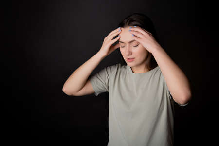 Headache. A young brunette in a T-shirt clutched her head with her hand, illness or fatigue. Portrait on a black backgroundの写真素材