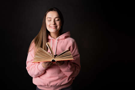 A young cute brunette in a pink hoodie, reading a book. Student with an open book in her hands, portrait on black. Study or read fictionの写真素材