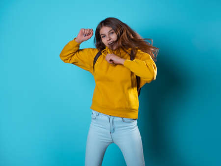 Beautiful young brunette in a yellow oversize hoodie having fun and jumping, portrait on a blue backgroundの写真素材