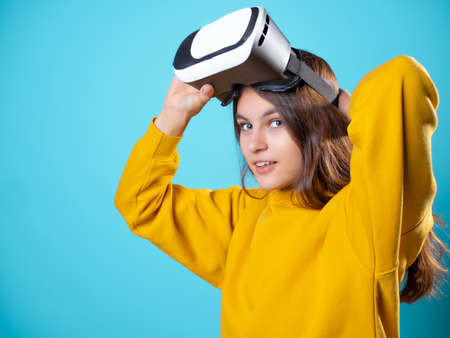 VR gamer, a young woman in a youth outfit uses a virtual reality helmet. A brunette in yellow on a blue background puts a vr gadget on her headの写真素材