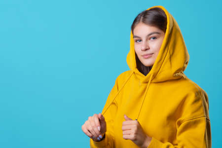 Beautiful young brunette with a hood on her head, youth oversize clothing style, portrait in a yellow hoodie on a blue background, bright colorsの写真素材
