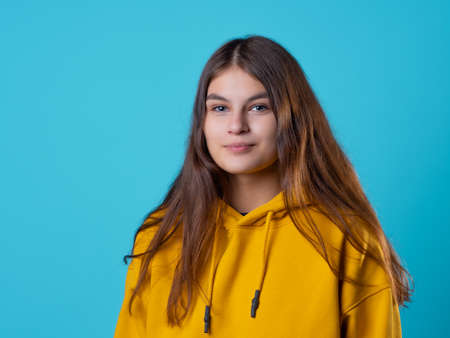 A beautiful young brunette in a yellow oversize hoodie, a positive student in a bright outfit, portrait on a blue backgroundの写真素材