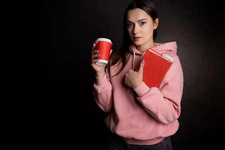 A young cute brunette in a pink hoodie, holding a book and coffee in her hands. Student on break, takeaway coffee, portrait on blackの写真素材