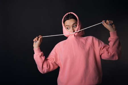 A cute young brunette in a pink hoodie hides her face comically pulling on a hood, portrait on a...