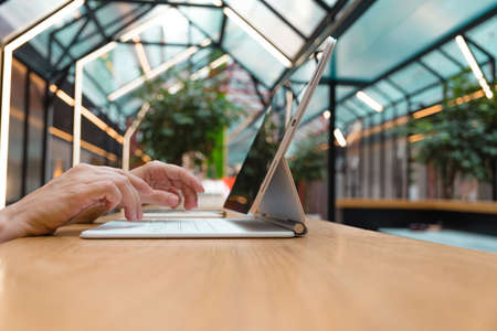 Remote work or freelance, modern opportunities for a mobile lifestyle. Womens hands are typing on keyboard of tablet computer, close-up side view, public space hall on background, low depth of field.の写真素材