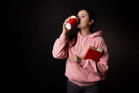 A young cute brunette in a pink hoodie, holding a book in her hands and drinking coffee. Student on break, portrait on blackの写真素材
