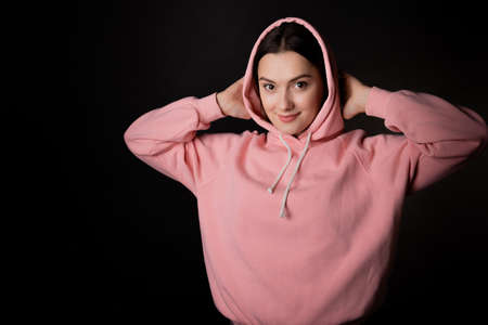 Cute young brunette in a pink hoodie puts a hood on her head, portrait on a black background.の写真素材