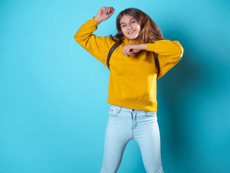 Beautiful young brunette in a yellow oversize hoodie having fun and jumping, portrait on a blue backgroundの写真素材
