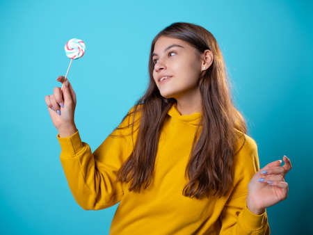 Beautiful young brunette with a lollipop in her hands, sweet candy, portrait in yellow on a blue background, bright colorsの写真素材