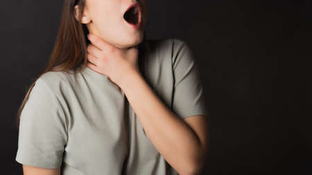 Sore throat, cold or virus, loss of voice. Psychological clamp. Young brunette in a T-shirt holding her throat, portrait on blackの写真素材