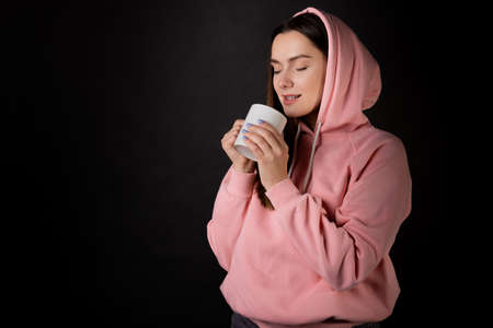 Cute young brunette in a pink hoodie drinking tea or coffee from a large mug, portrait on a black backgroundの写真素材