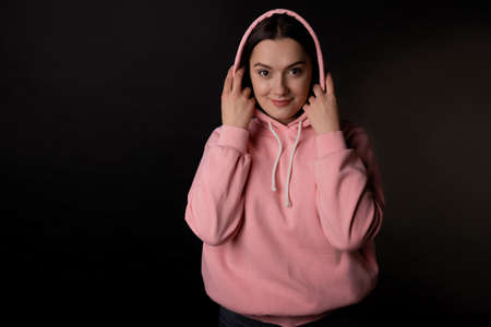Cute young brunette in a pink hoodie puts a hood on her head, portrait on a black background.の写真素材
