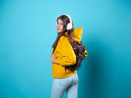 A young smiling student with a backpack and headphones, listening to music or recording a lecture. A young brunette in a bright yellow hoodie uses wireless headphonesの写真素材
