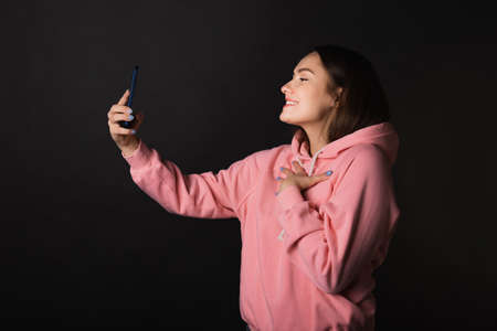 Cute young brunette in a pink hoodie uses a smartphone to take a selfie, portrait on a black backgroundの写真素材