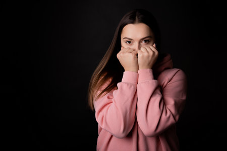 A cute young brunette in a pink hoodie hides her face with her hands, portrait on a black background.の写真素材