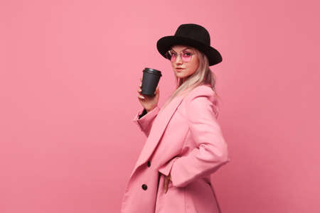Stylish trendy young woman in bright clothes on a pink background. Cool blonde girl in pink jacket and black hat with a coffee to take awayの写真素材