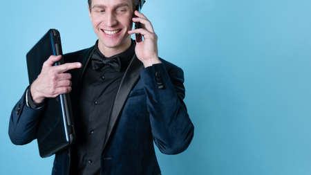 A successful and positive young man in a business suit with a laptop in his hands, talking on the phone, photo on blueの写真素材