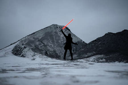 A fantastic assassin in a black suit with a red laser sword against a gray gloomy landscape of a snowy wasteland. dark photo with noise effectの写真素材