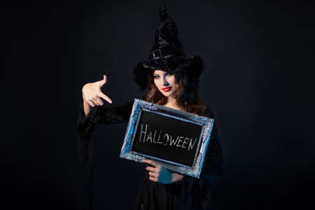 Halloween, a charming young witch in a costume with a pointed hat holds in her hands an inscription in a silver frame, the inscription Halloween in chalk on a black boardの写真素材