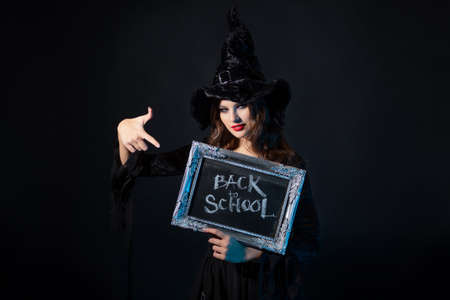 Magic school, a charming young witch in a suit with a pointed hat holding a silver-framed plaque with the inscription back to school, black chalkboard backgroundの写真素材