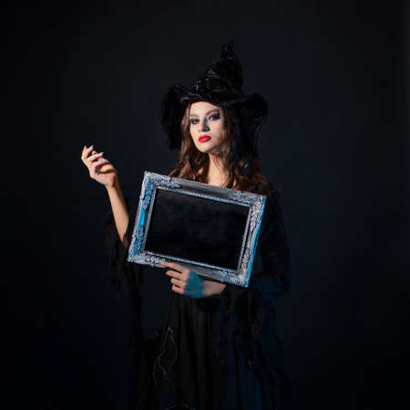 Halloween, a charming young witch in a costume with a pointed hat holding an empty plaque in a silver frame, a black chalkboard background, free space for your designの写真素材