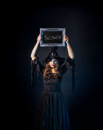 Halloween, a charming young witch in a costume with a pointed hat holds in her hands an inscription in a silver frame, the inscription Halloween in chalk on a black boardの写真素材