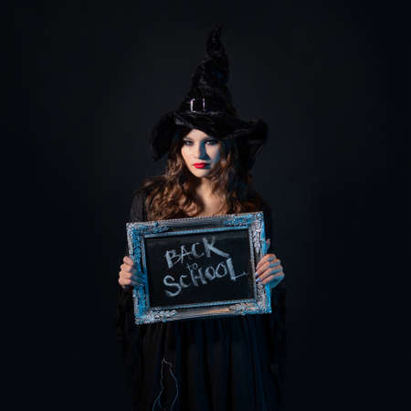 Magic school, a charming young witch in a suit with a pointed hat holding a silver-framed plaque with the inscription back to school, black chalkboard backgroundの写真素材