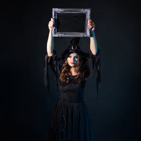 Halloween, a charming young witch in a costume with a pointed hat holding an empty plaque in a silver frame, a black chalkboard background, free space for your designの写真素材