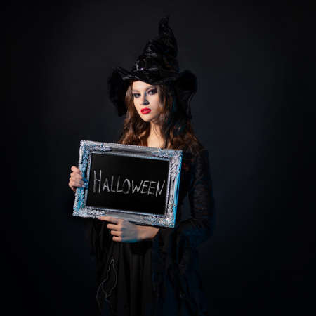 Halloween, a charming young witch in a costume with a pointed hat holds in her hands an inscription in a silver frame, the inscription Halloween in chalk on a black boardの写真素材