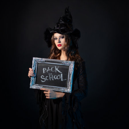 Magic school, a charming young witch in a suit with a pointed hat holding a silver-framed plaque with the inscription back to school, black chalkboard backgroundの写真素材