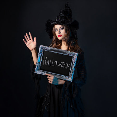 Halloween, a charming young witch in a costume with a pointed hat holds in her hands an inscription in a silver frame, the inscription Halloween in chalk on a black boardの写真素材