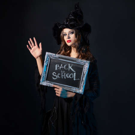 Magic school, a charming young witch in a suit with a pointed hat holding a silver-framed plaque with the inscription back to school, black chalkboard backgroundの写真素材