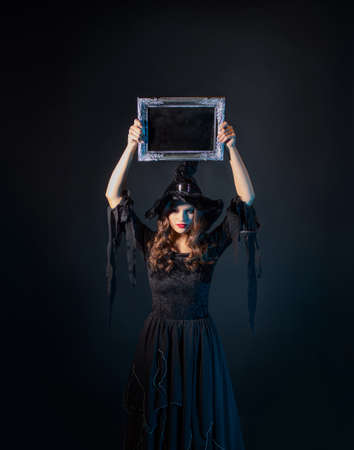 Halloween, a charming young witch in a costume with a pointed hat holding an empty plaque in a silver frame, a black chalkboard background, free space for your designの写真素材